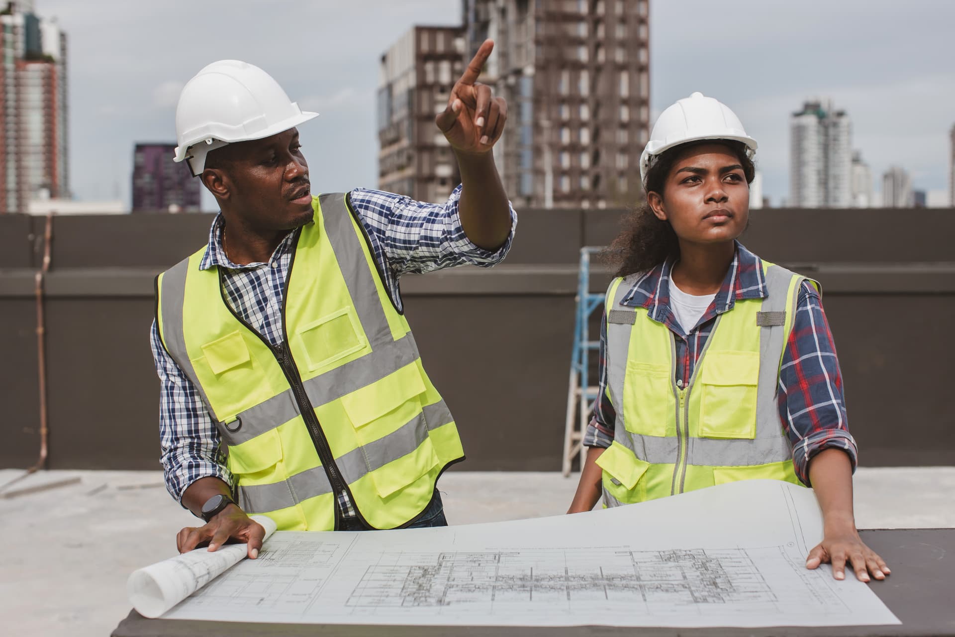 Civil engineers in hard hats and high-visibility vests reviewing blueprints on a construction site with a city skyline behind them